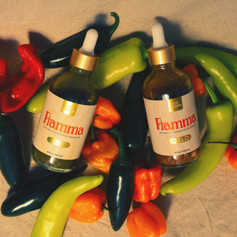 Two bottles of Fiamma pepper sauce surrounded by colorful peppers on a textured surface.