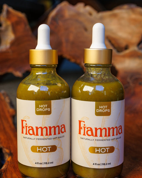 Two bottles of Fiamma hot sauce hot flavor with dropper lids on a gnarled wooden background.