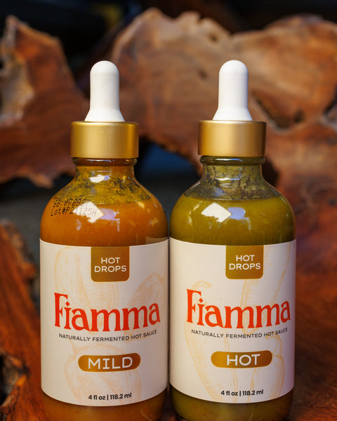 Two bottles of Fiamma hot sauce mild and hot flavor with dropper lids on a gnarled wooden background.