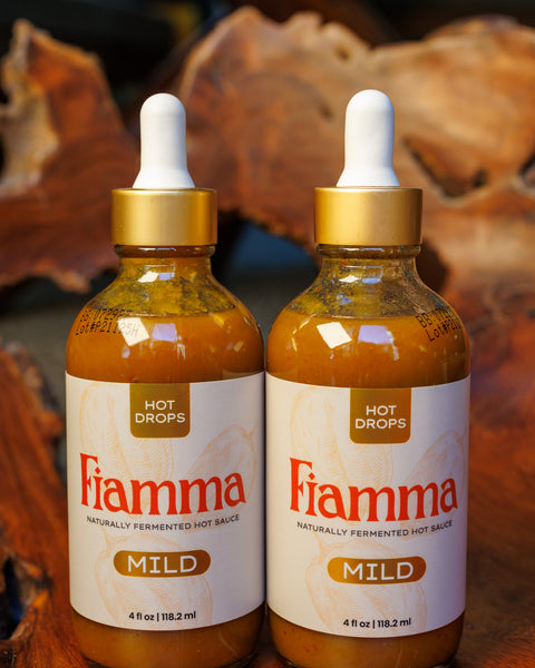 Two bottles of Fiamma hot sauce mild flavor with dropper lids on a gnarled wooden background.